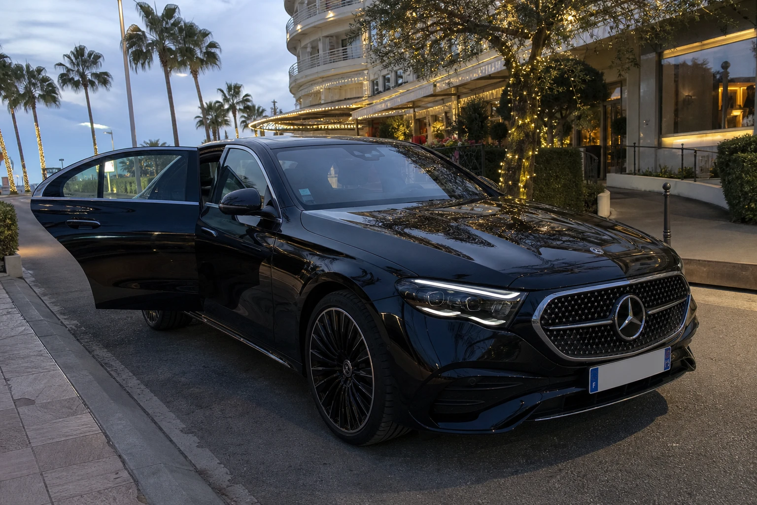 Mercedes Classe E noire, portière arrière ouverte, garée le soir sur La Croisette à Cannes près d'un hôtel de luxe illuminé — voiture de prestige pour soirée événementielle