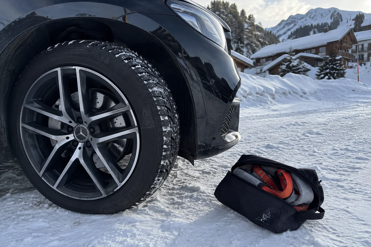 Roue d'une Mercedes noire équipée d'un pneu hiver certifié sur la neige d'une station de ski, chaussettes à neige posées à côté — conformité loi Montagne